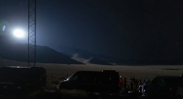 A mostly dark scene of vans and people in a desert area at night, but with a very bright sun-like light on the left sky.