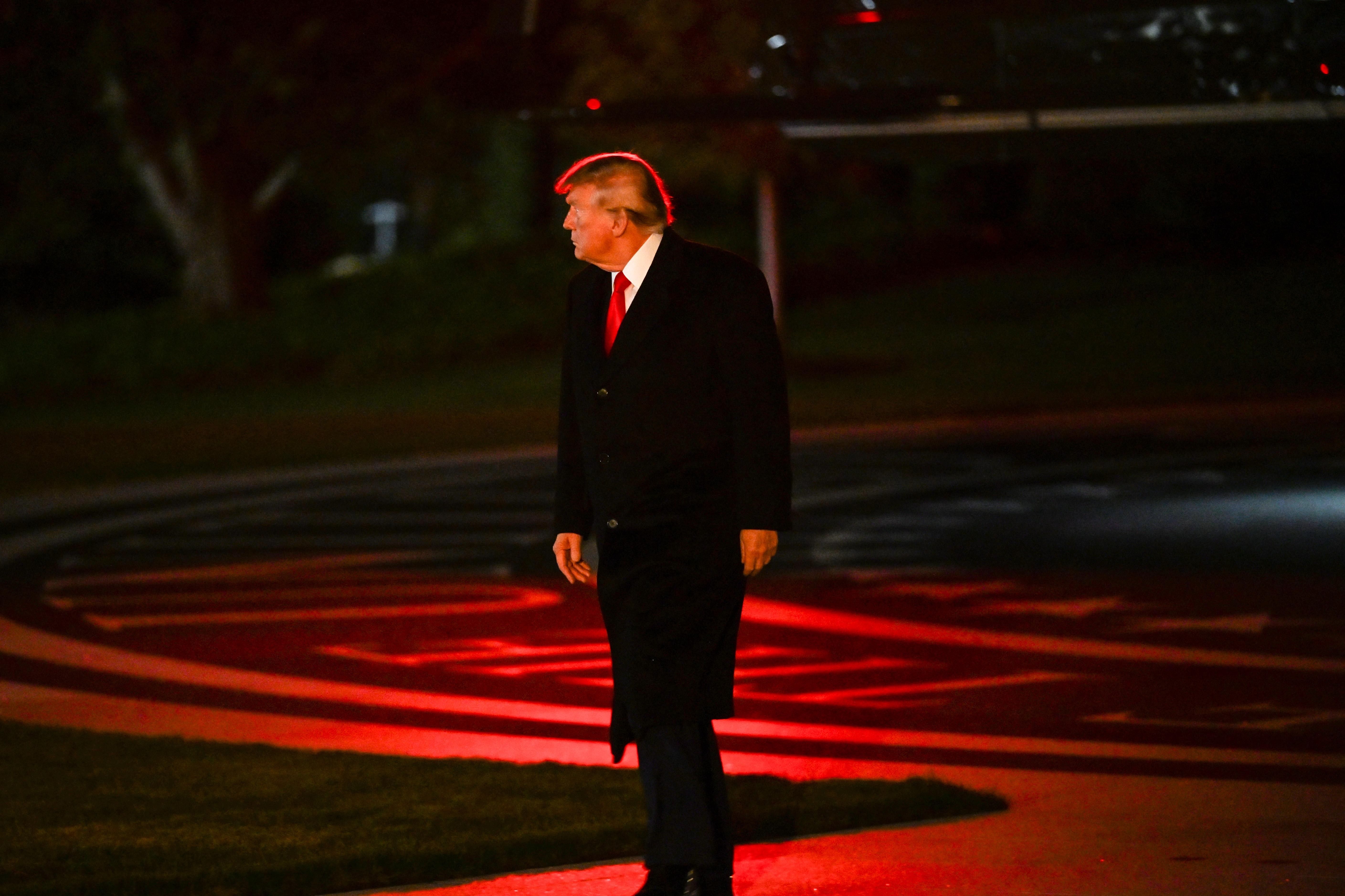 Donald Trump, lit with red light, walks away from the White House helipad on the South Lawn at night.