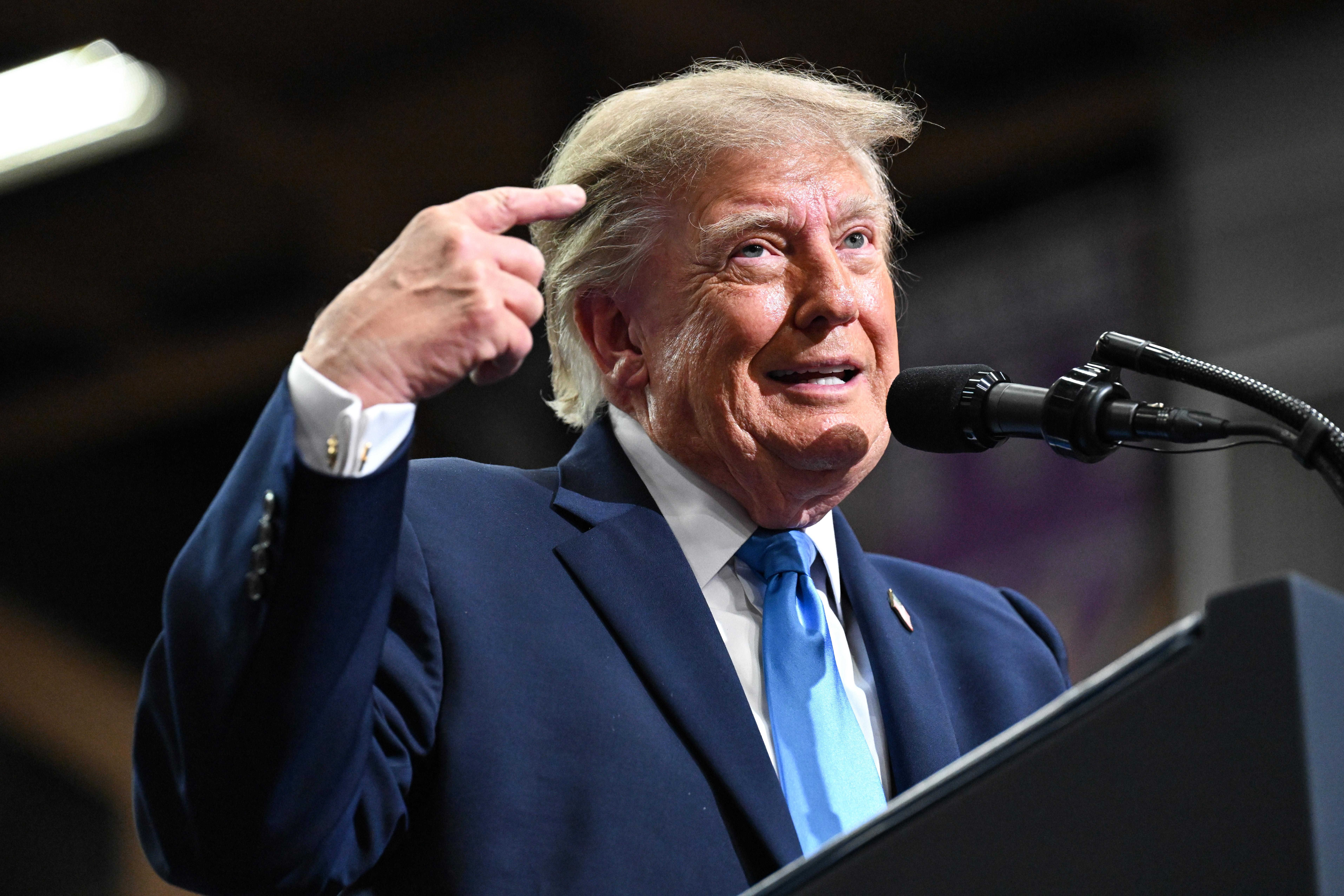 Donald Trump, wearing a suit and tie and standing on a stage behind a podium with a microphone, points at himself with his right hand.