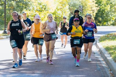 A runner in a yellow "Pacer" tank top jogs with a group of other runners.