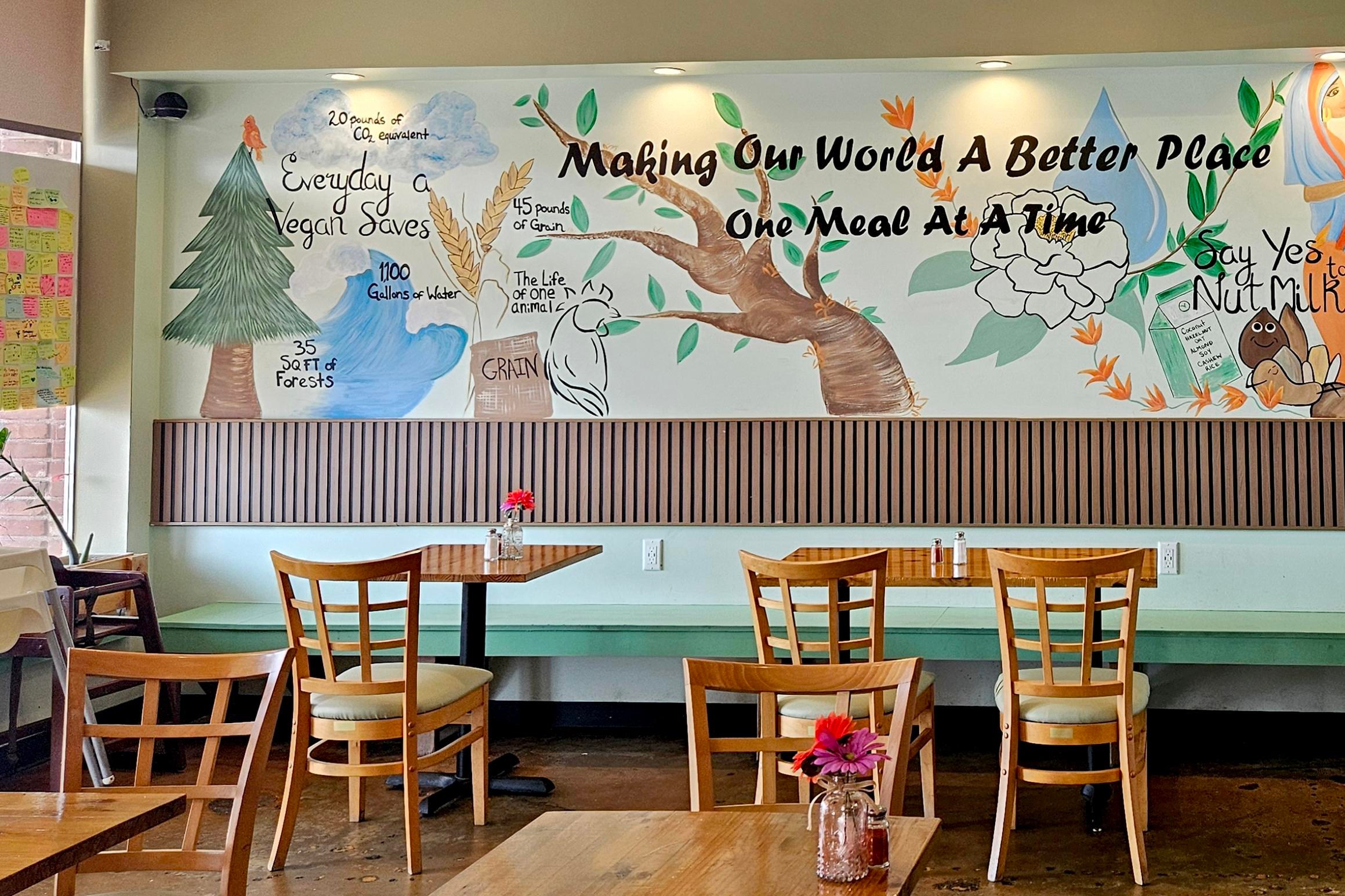 a painted mural on the wall of a cafe reads “Making our world a better place one meal at a time” and depicts various natural elements and food items. a painted mural on the wall of a cafe reads “Making our world a better place one meal at a time” and depicts various natural elements and food items.