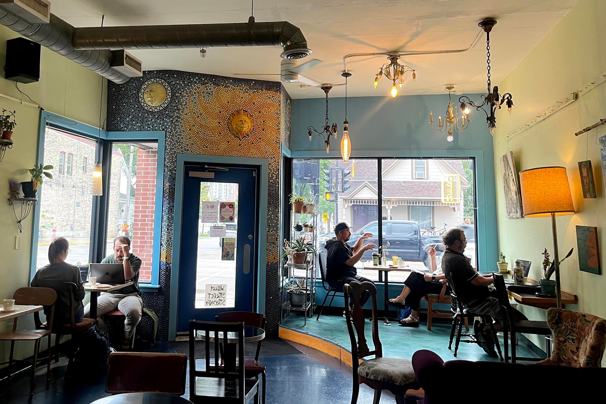 a blue, green, and yellow cafe interior with mosaic art and eclectic hanging lights. Several people are eating and working at small tables. a blue, green, and yellow cafe interior with mosaic art and eclectic hanging lights. Several people are eating and working at small tables.