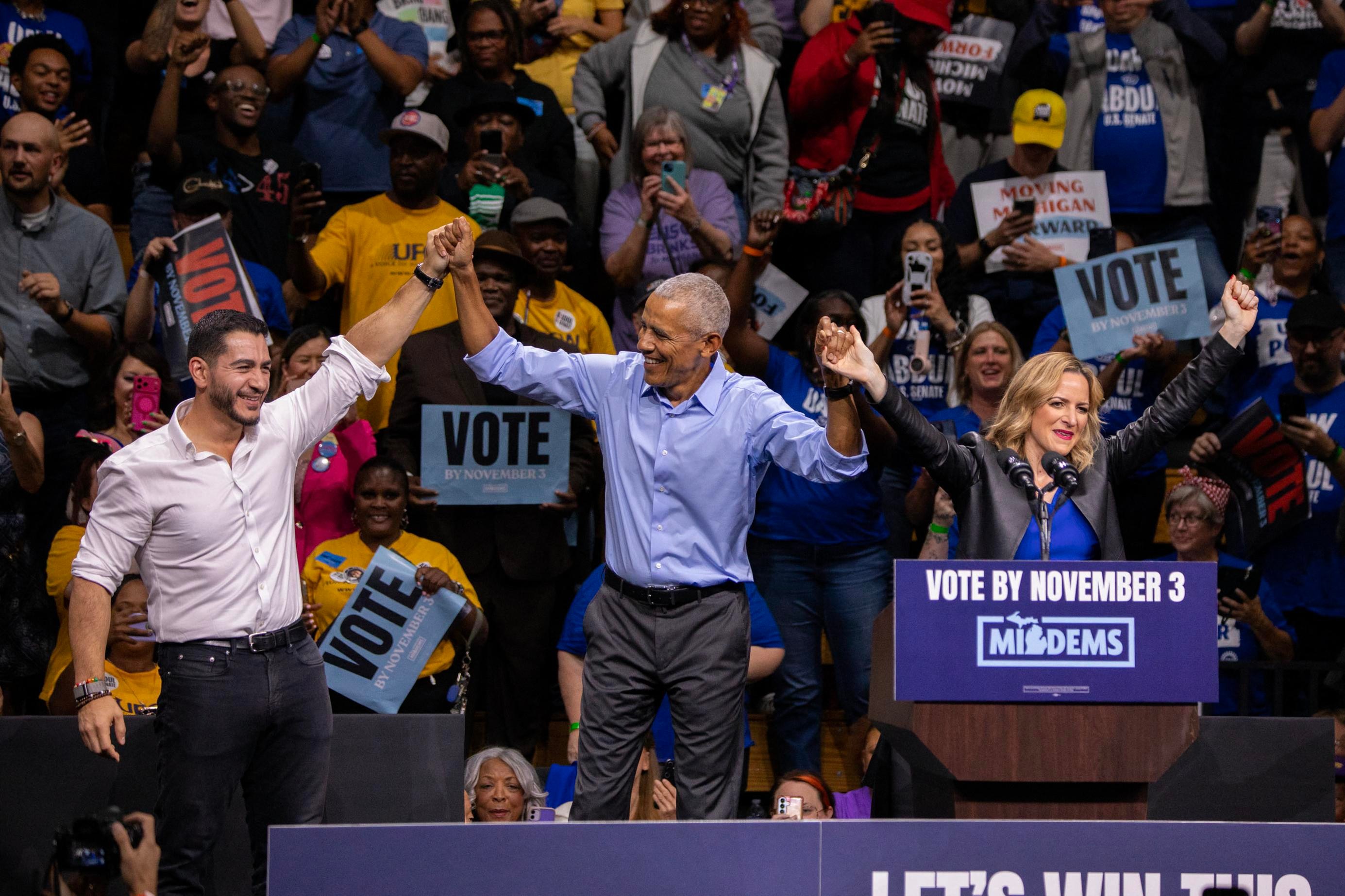 From left to right, Abdul El-Sayed, Barack Obama, and Jocelyn Benson hold raised hands and smile in front of a crowd of supporters. 