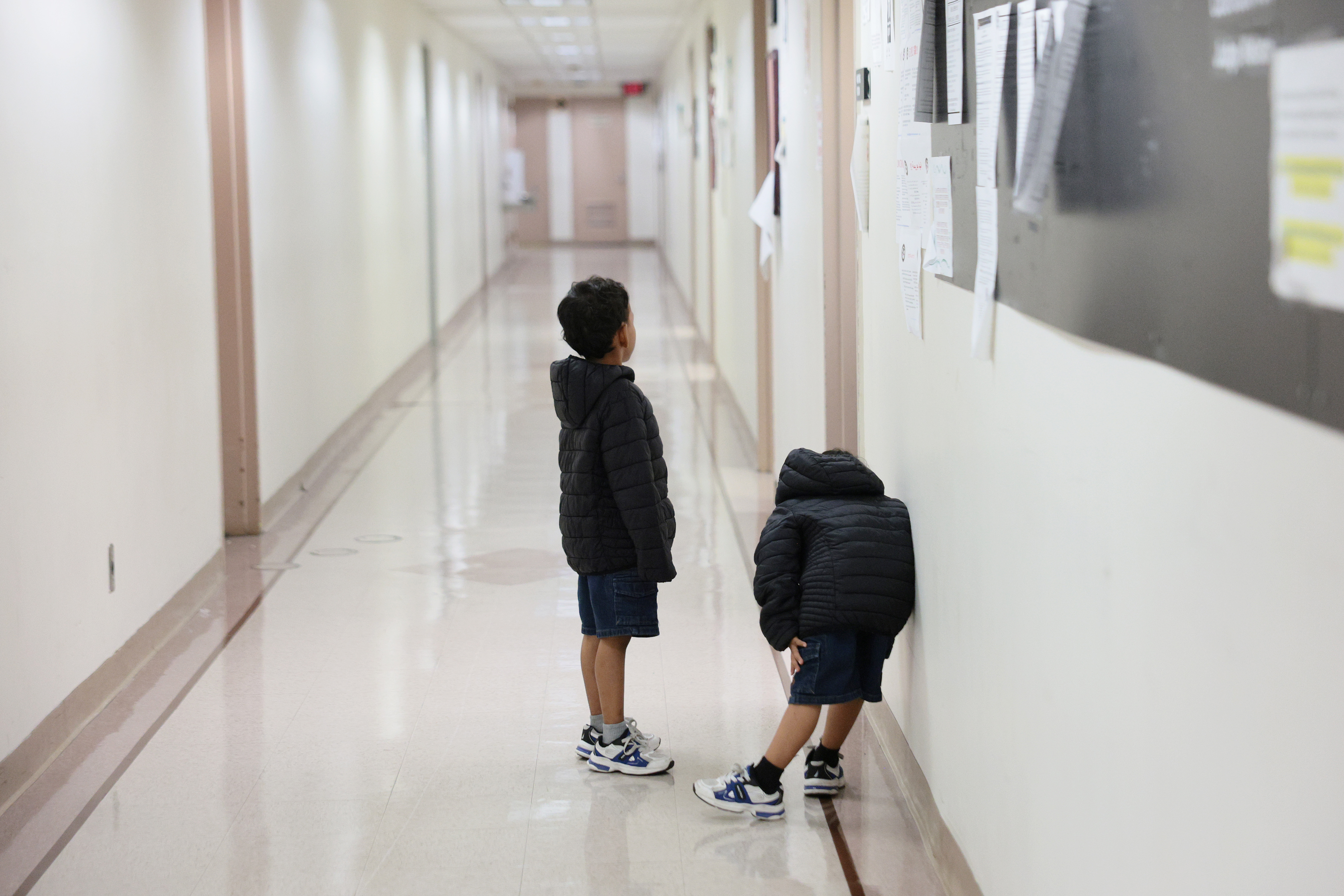 Two children stand alone in a long white hallway. 