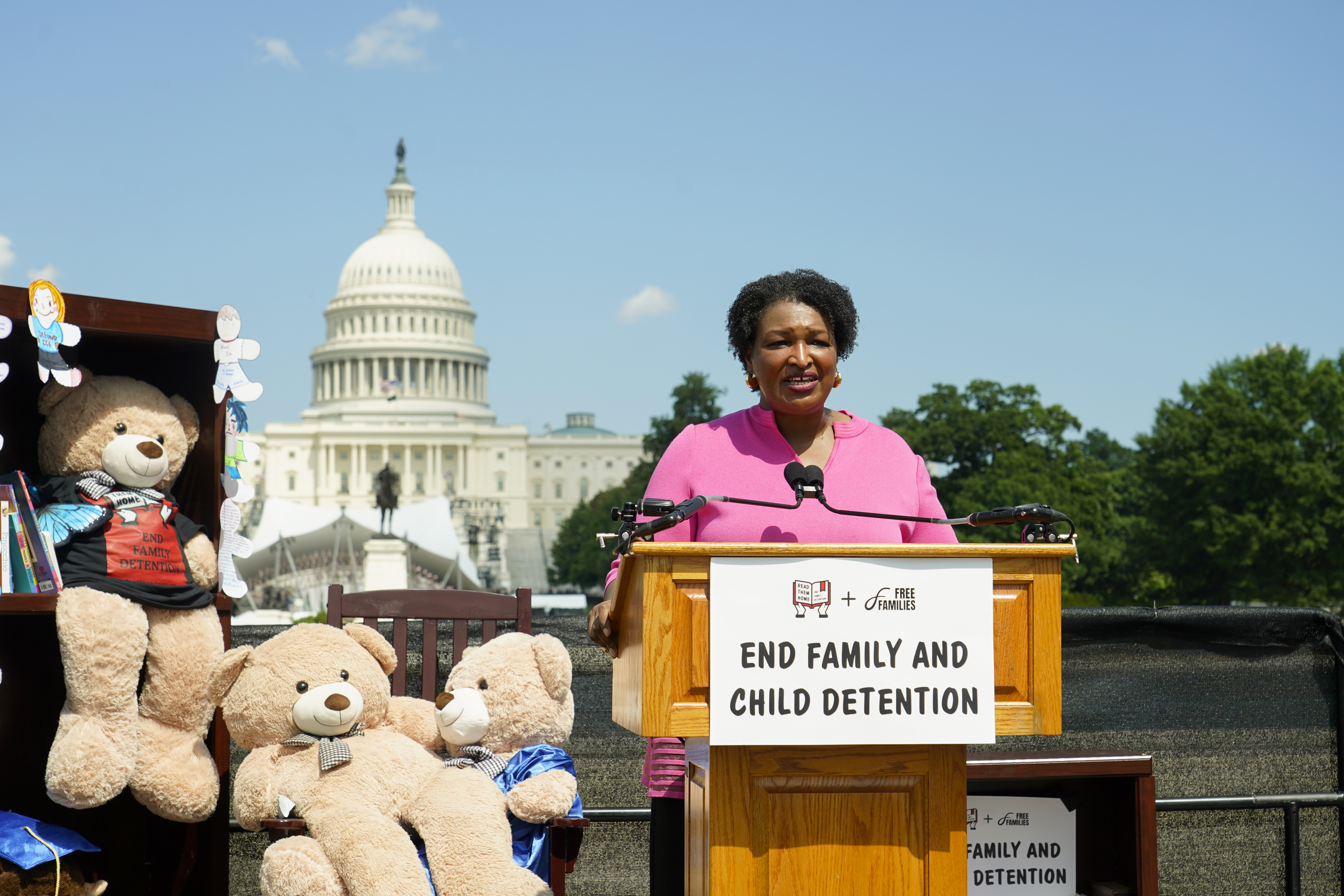 Stacey Abrams speaks at a podium next to large stuffed teddy bears, with the US Capitol building behind her.